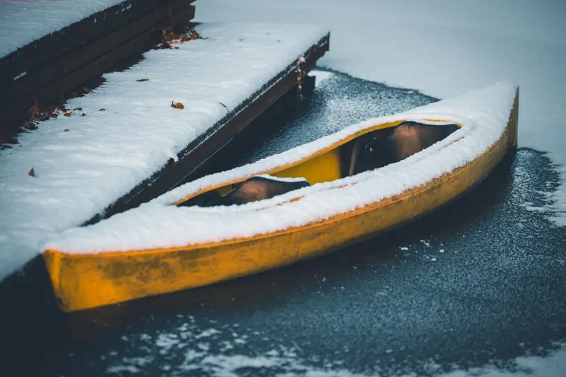 A snow-covered yellow kayak sits on a partially frozen lake next to a wooden dock in winter