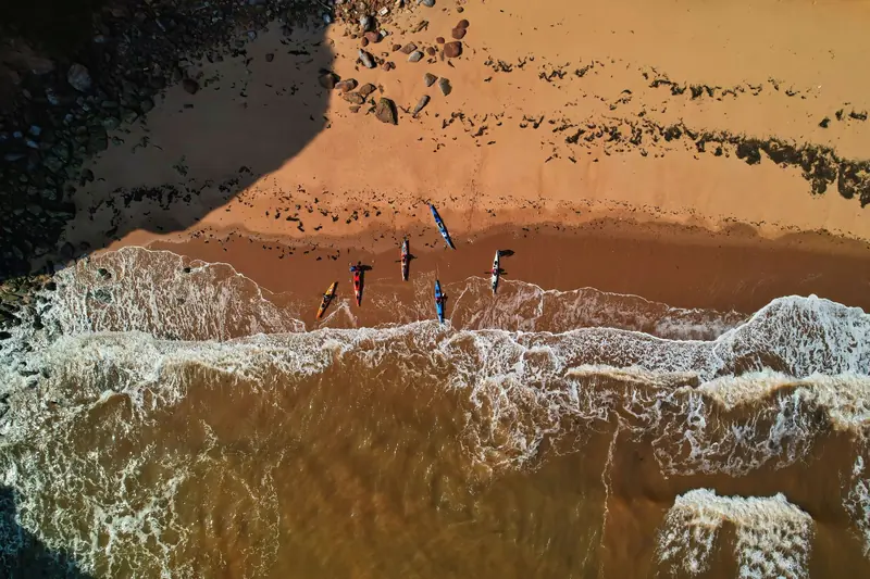 Drone shot capturing kayaks lined on a sandy shore of the UK coast, with waves approaching