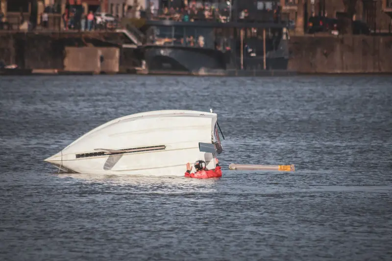 A capsized boat near an urban waterfront with a person wearing a life jacket visible in calm waters