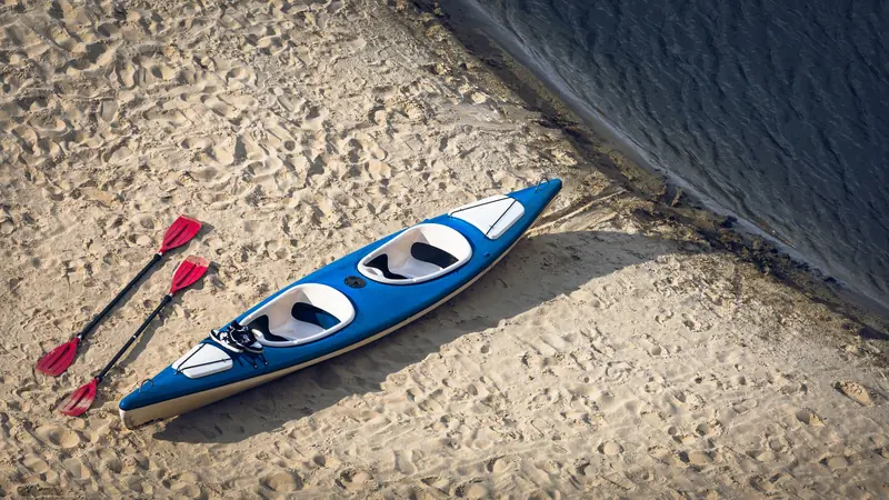 A solitary blue kayak and two red paddles rest on a sandy beach by the water's edge
