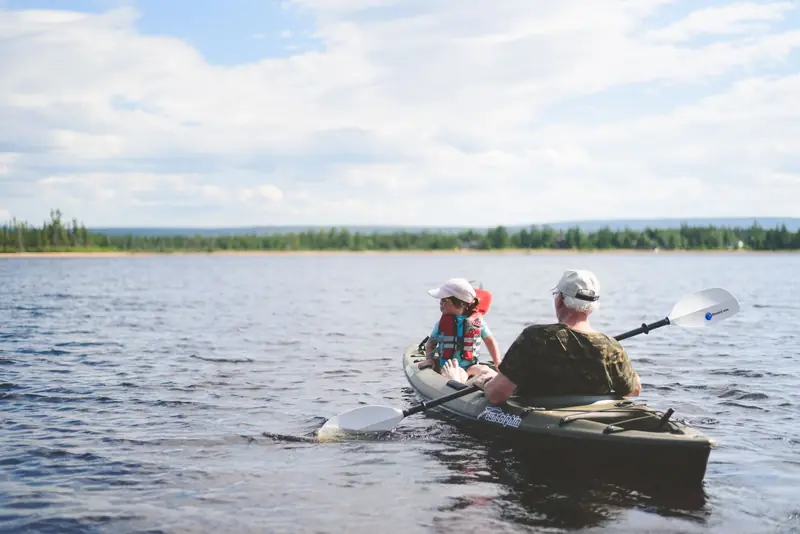 A grandfather and grandson kayaking together on a serene lake under a blue sky, enjoying outdoor adventure