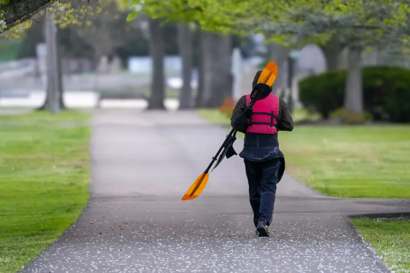 An individual walks through a park carrying kayak paddles, wearing a pink life jacket